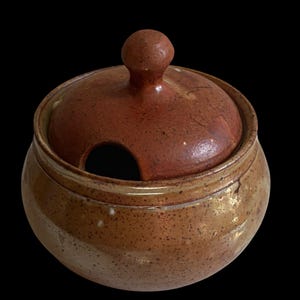 May include: A round ceramic sugar bowl with a speckled tan and brown glaze. The bowl features a matching lid with a central knob handle and a small semi-circular cutout on the rim to accommodate a spoon, showcasing a textured finish.