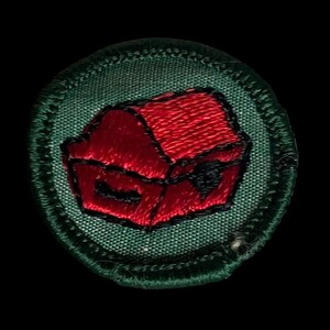May include: Circular embroidered patch depicting a red treasure chest with a black lock, on a green background. The patch has a green border with detailed stitching. The design is suitable for sewing onto fabric, with a textured appearance.