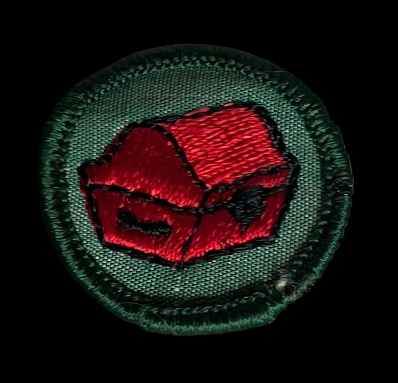 May include: Circular embroidered patch depicting a red treasure chest with a black lock, on a green background. The patch has a green border with detailed stitching. The design is suitable for sewing onto fabric, with a textured appearance.