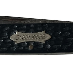 May include: A folding pocket knife with a black textured handle featuring a silver-toned metal inlay plate engraved with the words SCOUT KNIFE. The knife has metallic end caps and a small circular hole on the left side of the handle casing.