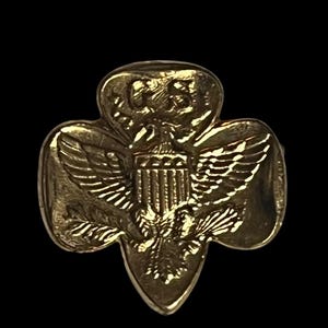 May include: A gold-coloured pin in the shape of a trefoil, featuring a detailed depiction of an eagle with outstretched wings, a shield, and the letters "US". The pin has a shiny, metallic finish.