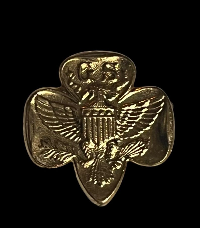 May include: A gold-coloured pin in the shape of a trefoil, featuring a detailed depiction of an eagle with outstretched wings, a shield, and the letters "US". The pin has a shiny, metallic finish.