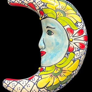May include: A crescent moon wall hanging made of ceramic, featuring a hand-painted face with light blue skin and red lips. The outer edge is decorated with a blue and white grid pattern, while the inner curve displays yellow and green floral motifs.
