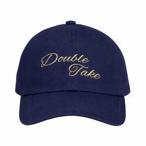 May include: Navy blue baseball cap with the embroidered text "Double Take" in a gold script font. The cap has a curved brim and a button on top. A classic, casual accessory.