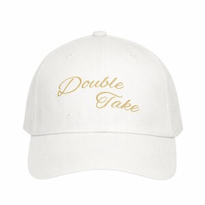 May include: White baseball cap with a curved brim. The cap features the text "Double Take" embroidered in a gold script font across the front. The hat is a classic style, suitable for casual wear.