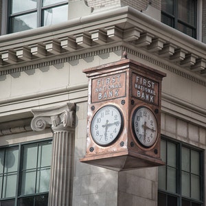May include: A copper-colored clock with two faces, each displaying the time with Roman numerals. The clock is mounted on a building and has the text "FIRST NATIONAL BANK" on each side.