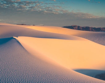 May include: A landscape photograph of rolling white sand dunes under a blue and cloudy sky. The dunes are illuminated by warm sunlight, creating shadows and highlighting the textured patterns on the sand.