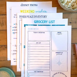 May include: Several white paper sheets with printed titles such as "Dinner Menu" and "Grocery List." The sheets feature blue and teal accents, with handwritten notes and a pen. The words "Weekend Wishes" are also visible.