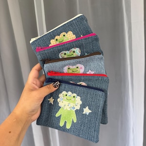May include: A stack of four denim zipper pouches, each featuring a light green frog appliqué with a patterned fabric collar. The pouches have zippers in white, pink, brown, and navy blue, with small fabric star accents on the front of the pouches.