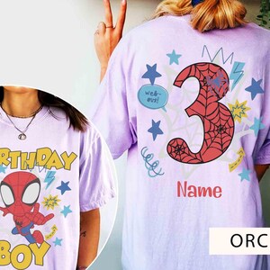 Pu&ograve; includere: Una t-shirt color orchidea con la grafica di un supereroe rosso e blu sul davanti con il testo "BIRTHDAY BOY" e "web-out!". Il retro mostra un grande numero 3 con motivo a ragnatela rossa, la parola "Name" e delle stelle.