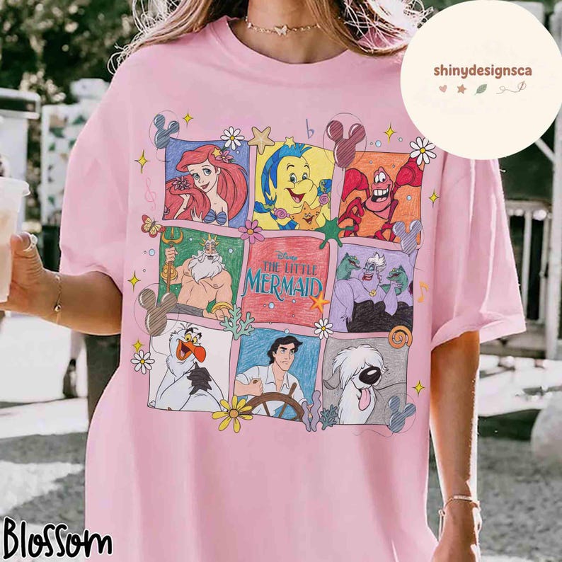 May include: Pink t-shirt featuring a colorful graphic of characters from The Little Mermaid. The design includes Ariel, Flounder, Sebastian, King Triton, Ursula, Prince Eric, and Max. The shirt has short sleeves and a crew neck.