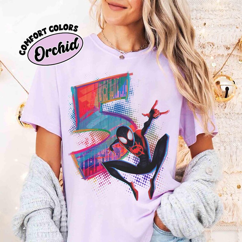 May include: A lilac t-shirt with a vibrant graphic of the number 5 and a black and red Spider-Man character. The shirt has the words "COMFORT COLORS" and "Orchid" printed on it.
