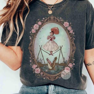 Puede incluir: Una camiseta gris carb&oacute;n con un marco ovalado central decorado con rosas rosadas. Dentro del marco, una persona con vestido blanco sostiene una sombrilla rosa y camina sobre una cuerda floja sobre un cocodrilo con la boca abierta.