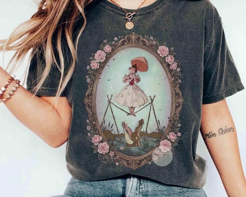 Puede incluir: Una camiseta gris carb&oacute;n con un marco ovalado central decorado con rosas rosadas. Dentro del marco, una persona con vestido blanco sostiene una sombrilla rosa y camina sobre una cuerda floja sobre un cocodrilo con la boca abierta.