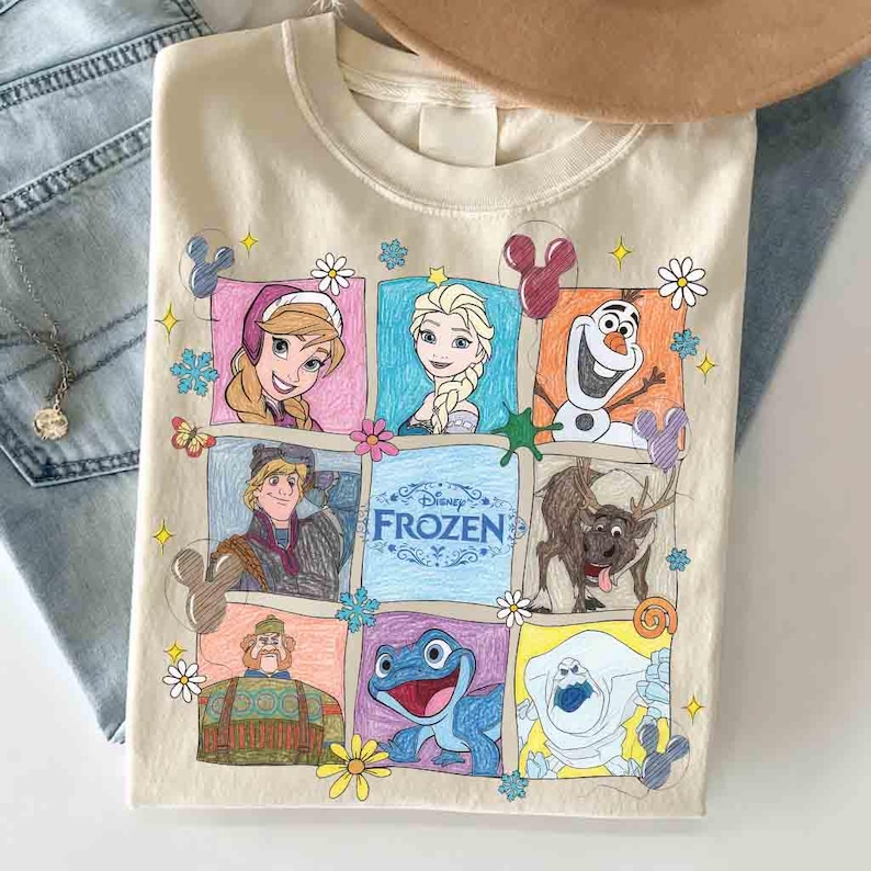 Op de afbeelding: Beige crewneck sweatshirt met een kleurrijke afbeelding van personages uit de Disney-film "Frozen". Het ontwerp bevat Anna, Elsa, Olaf, Kristoff, Sven en andere personages. Het woord "Frozen" wordt ook weergegeven.