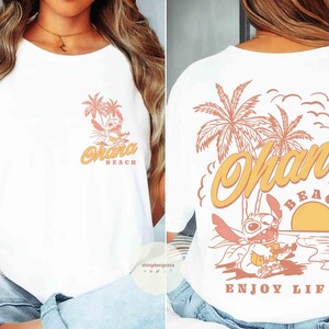 May include: White t-shirt with a front and back design. The front features a cartoon character with palm trees and the words "Ohana Beach". The back has a larger design with the same character, palm trees, a sunset, and the words "Ohana Beach Enjoy Life".