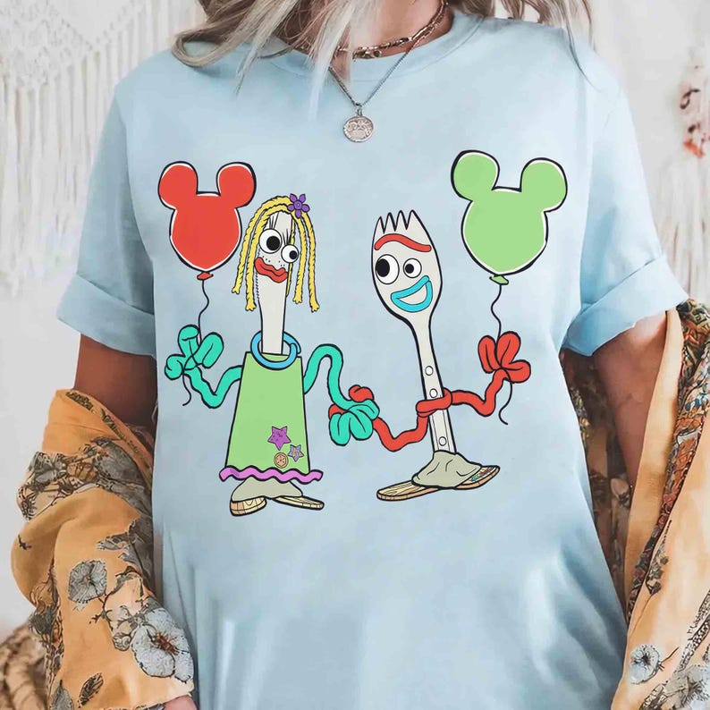 May include: Light blue t-shirt featuring a cartoon illustration of two anthropomorphic utensils holding Mickey Mouse-shaped balloons. One utensil is a fork wearing a green dress, and the other is a spoon. The balloons are red and green.
