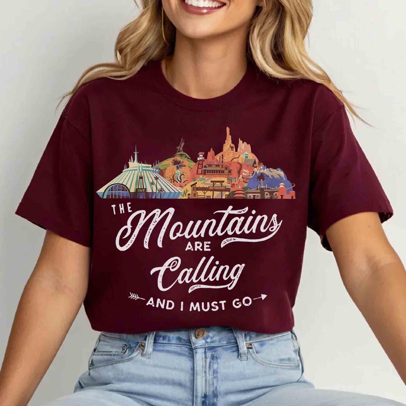 May include: A maroon short-sleeve t-shirt featuring a graphic of various mountain peaks and structures. Below the graphic, white text reads: THE Mountains ARE Calling AND I MUST GO, with a small arrow icon pointing to the left and right.