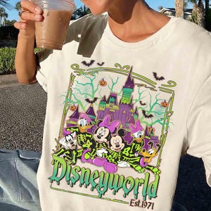 May include: Cream-coloured t-shirt with a Halloween-themed Disney World graphic. The design features cartoon characters in skeleton costumes, a purple castle, bats, and pumpkins. The text "Disneyworld" and "Est. 1971" are also visible.