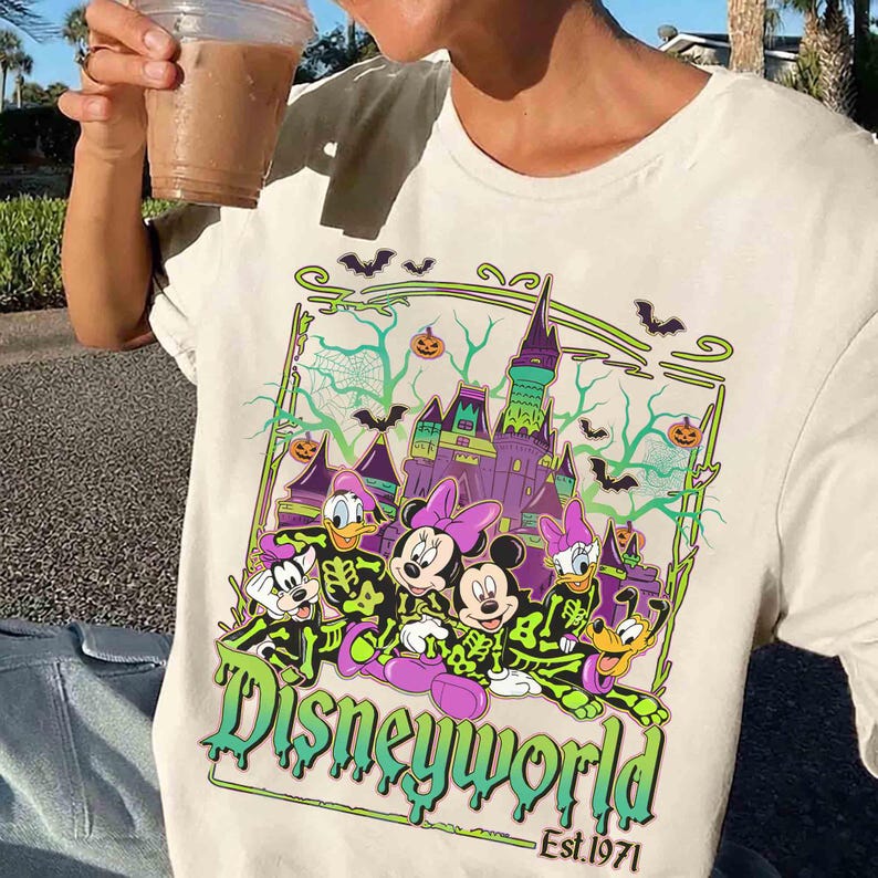 May include: Cream-coloured t-shirt with a Halloween-themed Disney World graphic. The design features cartoon characters in skeleton costumes, a purple castle, bats, and pumpkins. The text "Disneyworld" and "Est. 1971" are also visible.