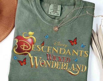 May include: An olive green short-sleeve t-shirt featuring a graphic of a red apple and the text Disney Descendants Wicked Wonderland in gold lettering. The design includes several small orange and blue butterflies scattered across the front.