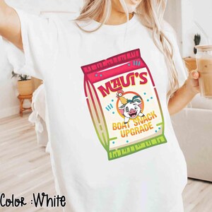 Puede incluir: Camiseta blanca con un gr&aacute;fico colorido de una bolsa de snacks con el texto "Maui's Boat Snack Upgrade" y un cerdo y un gallo de dibujos animados. El gr&aacute;fico utiliza tonos de rojo, rosa, verde y beige.