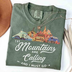 May include: An olive green t-shirt featuring a graphic of various mountain-themed attractions and the white text "THE Mountains ARE Calling AND I MUST GO" with arrow accents. The shirt has a crew neckline and a Comfort Colors brand label.