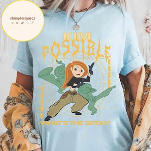 May include: Light blue t-shirt featuring a cartoon graphic of Kim Possible with the text "KIM POSSIBLE" in yellow and the phrase "WHAT'S THE SITCH?" in white. The design includes green silhouettes of Kim Possible in action poses.