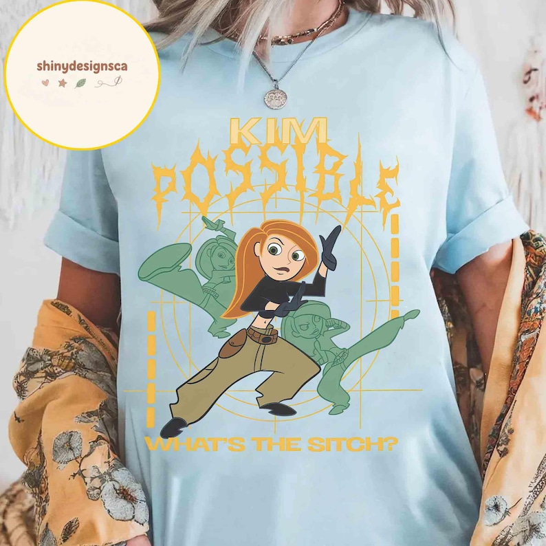 May include: Light blue t-shirt featuring a cartoon graphic of Kim Possible with the text "KIM POSSIBLE" in yellow and the phrase "WHAT'S THE SITCH?" in white. The design includes green silhouettes of Kim Possible in action poses.
