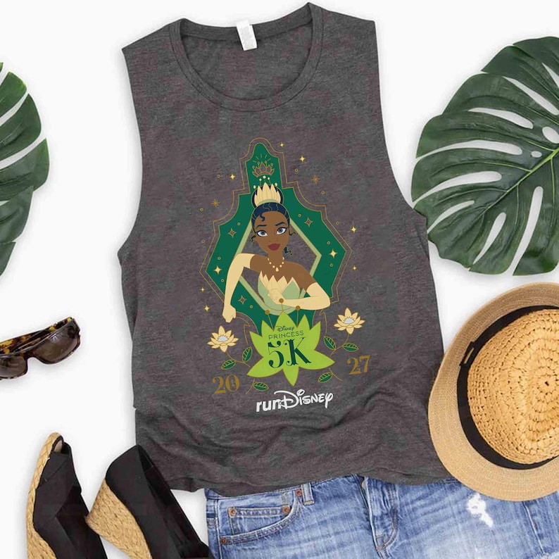 May include: A dark gray sleeveless tank top featuring a graphic of Princess Tiana from The Princess and the Frog. The design includes a green leaf shape with the text 5K, the numbers 20 and 27, and the runDisney logo printed in white ink.