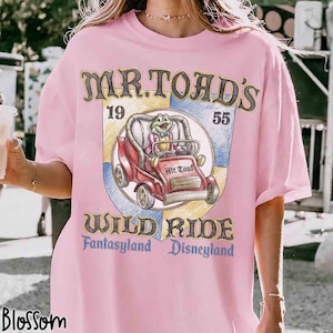 K&ouml;nnte beinhalten: Hellrosa T-Shirt mit einem Aufdruck von Mr. Toad's Wild Ride aus Disneyland. Das Design zeigt einen Cartoon-Frosch in einem roten Auto, mit dem Text "Mr. Toad's Wild Ride, Fantasyland, Disneyland."