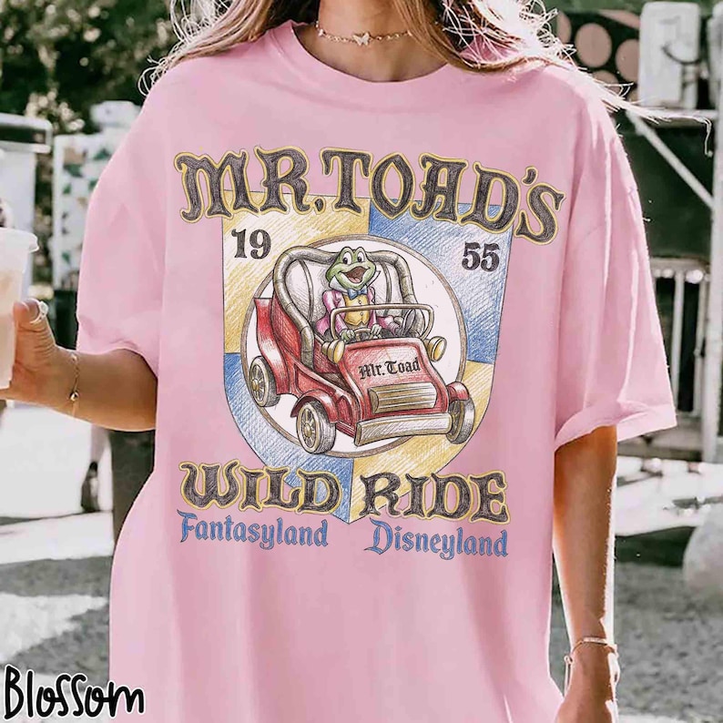 K&ouml;nnte beinhalten: Hellrosa T-Shirt mit einem Aufdruck von Mr. Toad's Wild Ride aus Disneyland. Das Design zeigt einen Cartoon-Frosch in einem roten Auto, mit dem Text "Mr. Toad's Wild Ride, Fantasyland, Disneyland."