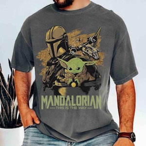 Puede incluir: Camiseta gris oscuro con un dise&ntilde;o de The Mandalorian, Grogu y una nave espacial. El texto "MANDALORIAN - THIS IS THE WAY" est&aacute; impreso en verde. El dise&ntilde;o tiene un aspecto vintage y desgastado.
