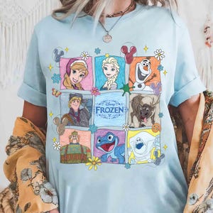 Op de afbeelding: Lichtblauw T-shirt met een kleurrijke afbeelding van personages uit de Disney-film "Frozen". Het ontwerp bevat afbeeldingen van Anna, Elsa, Olaf, Kristoff, Sven en andere personages, geplaatst in een raster van kleurrijke vierkanten. Het woord "Frozen" is ook zichtbaar.