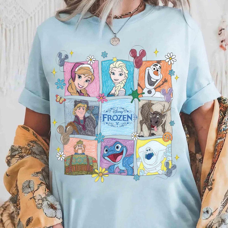 Op de afbeelding: Lichtblauw T-shirt met een kleurrijke afbeelding van personages uit de Disney-film "Frozen". Het ontwerp bevat afbeeldingen van Anna, Elsa, Olaf, Kristoff, Sven en andere personages, geplaatst in een raster van kleurrijke vierkanten. Het woord "Frozen" is ook zichtbaar.