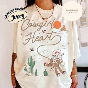 May include: Ivory-colored t-shirt with the words "Cowgirl at Heart" in a rope-like font. The design includes a cartoon cowgirl riding a horse, a desert landscape, and the Pixar logo.