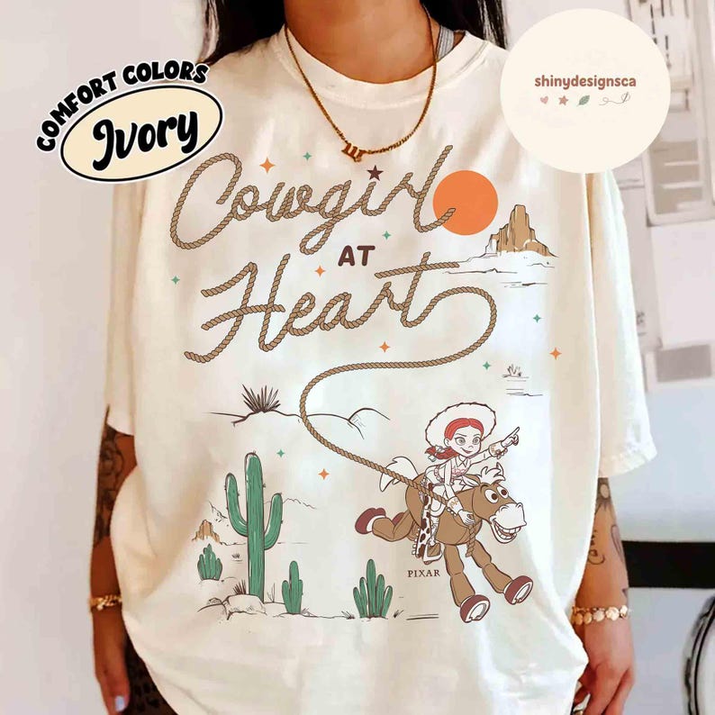 May include: Ivory-colored t-shirt with the words "Cowgirl at Heart" in a rope-like font. The design includes a cartoon cowgirl riding a horse, a desert landscape, and the Pixar logo.