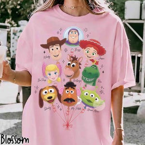 Op de afbeelding: Een lichtroze t-shirt met een grafische print van verschillende Toy Story-personagehoofden ontworpen als ballonnen. De personages zijn onder andere Woody, Buzz, Jessie, Bo Peep, Bullseye, Rex, Slinky, Mr. Potato Head en een groene Alien, verbonden door rode lijnen.