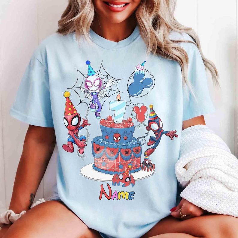 May include: Light blue t-shirt featuring a birthday cake with a number 7 candle, Spider-Man characters, and the word "Name" in colorful letters. The design includes balloons and a spider web.