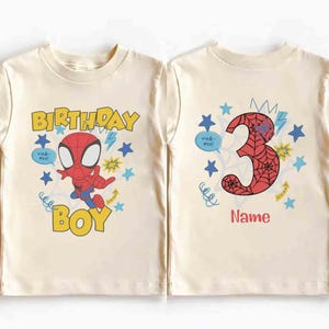 Pu&ograve; includere: Due magliette color crema. La maglietta a sinistra presenta un personaggio ragno dei cartoni animati, il testo "BIRTHDAY BOY" e stelle blu. Quella a destra mostra un grande numero "3" con motivo a ragnatela, stelle blu e il testo "Name".