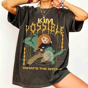 以下が含まれることがあります： チャコールグレーの半袖Tシャツで、アニメキャラクターのキム・ポッシブルがダイナミックなポーズをとっているグラフィックが描かれています。デザインには黄色の文字で「KIM POSSIBLE」と「WHAT'S THE SITCH?」と記され、円形のグリッド背景が配置されています。