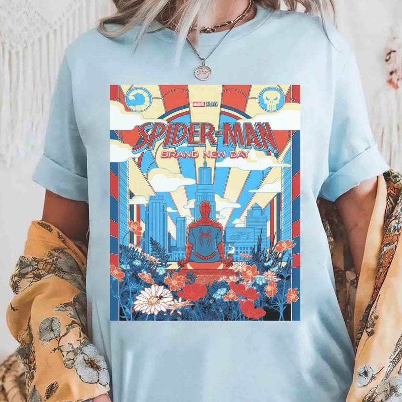 May include: A light blue t-shirt featuring a graphic print of Spider-Man sitting on a rooftop overlooking a city skyline. The design includes the text "SPIDER-MAN BRAND NEW DAY" and "MARVEL STUDIOS" with colorful flowers in the foreground.