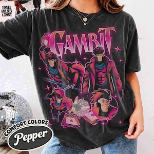 May include: A charcoal gray short-sleeve t-shirt featuring a graphic print of the comic book character Gambit in various action poses. The design includes the text GAMBIT in large pink letters, small pink star accents, and playing cards.