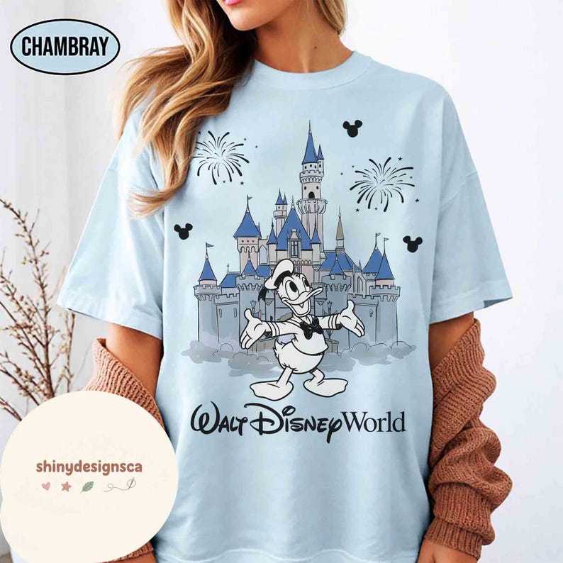 May include: Light blue t-shirt featuring a graphic of Donald Duck in front of Cinderella's castle at Walt Disney World. The design includes fireworks and Mickey Mouse silhouettes. The text "Walt Disney World" is printed below the castle.