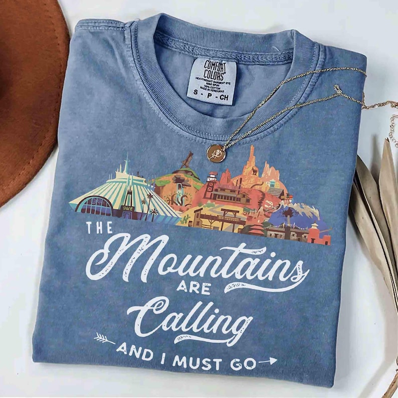 May include: A blue short-sleeve t-shirt with a graphic print of stylized mountain structures and the white text "THE Mountains ARE Calling AND I MUST GO". The shirt features a crew neckline and a Comfort Colors brand label on the inner collar.