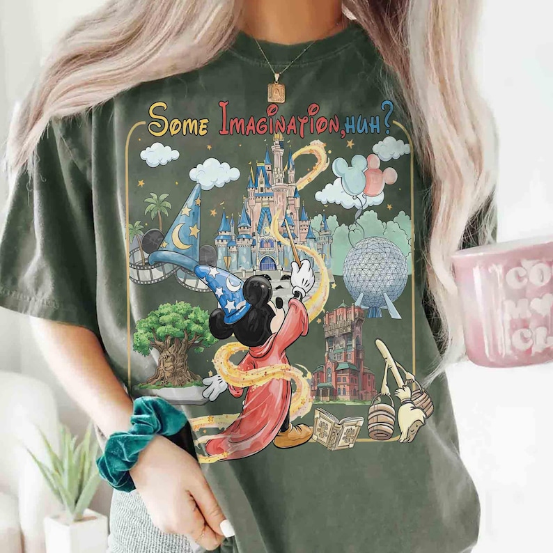 Op de afbeelding: Olijfgroen T-shirt met een grillige illustratie van Mickey Mouse als tovenaar, toverend voor een kasteel. Het shirt bevat de tekst "Some Imagination, Huh?" en andere iconische themaparkafbeeldingen.