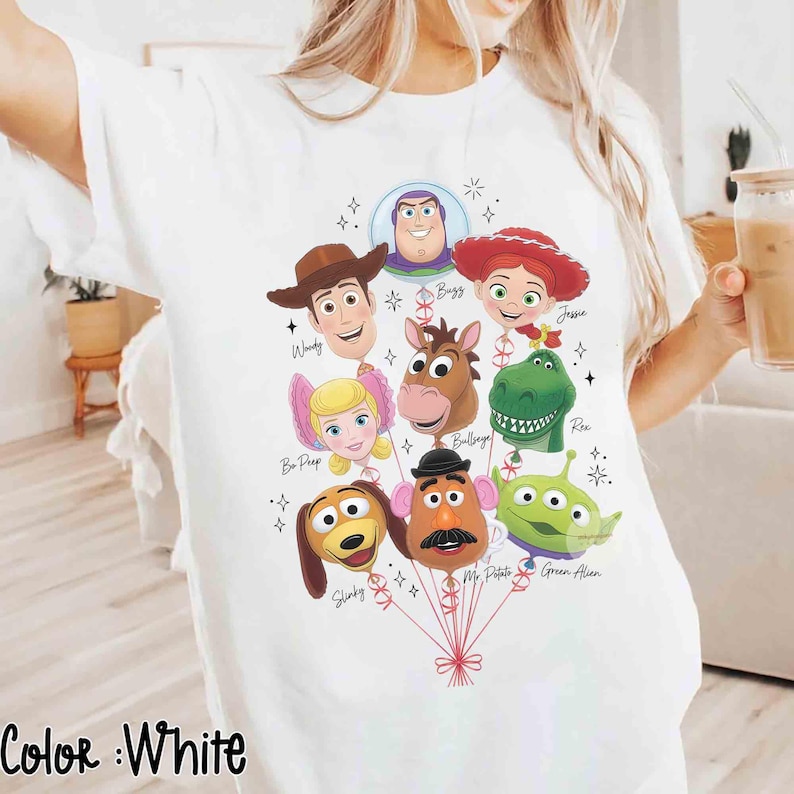 Op de afbeelding: Een wit t-shirt met een grafische print van Toy Story-personages ontworpen als ballonnen. De personages zijn onder andere Woody, Buzz Lightyear, Jessie, Bo Peep, Bullseye, Rex, Slinky Dog, Mr. Potato Head en een Alien, elk met hun naam.