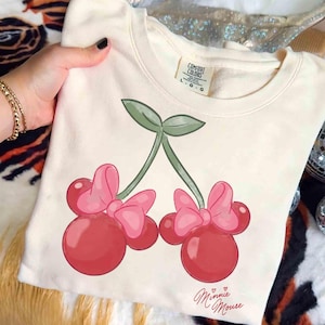 May include: Cream-colored sweatshirt featuring a Minnie Mouse cherry design. The graphic includes two red cherries with pink bows and green stems. The words "Minnie Mouse" are written in cursive below the cherries.