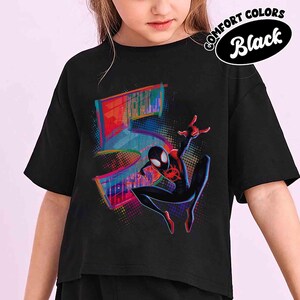 May include: Black t-shirt with a superhero graphic and the number "5" in a colourful, pixelated design. The words "COMFORT COLORS Black" are in a circular design at the top. The superhero is in a dynamic pose.