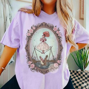 Puede incluir: Una camiseta de color morado claro con un marco ovalado central decorado con rosas rosas. Dentro del marco, una ilustraci&oacute;n muestra a una figura con vestido y sombrilla haciendo equilibrio sobre una cuerda sobre un caim&aacute;n marr&oacute;n en el agua.